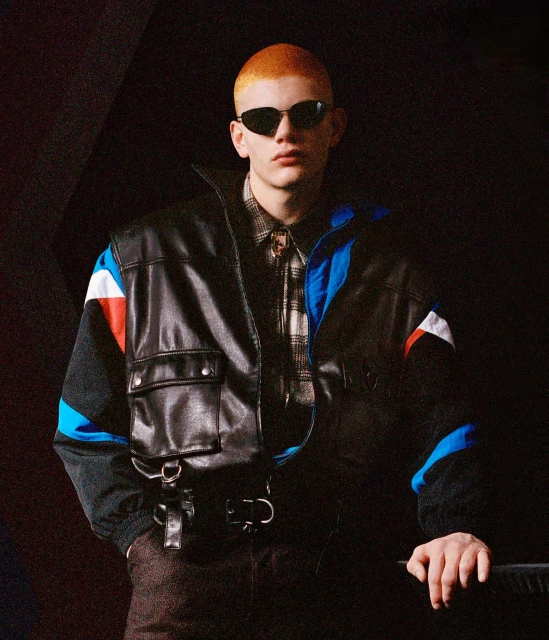 An adult male with a shaved ginger head wearing a dark leather bomber jacket and sunglasses poses in front of a dark background.