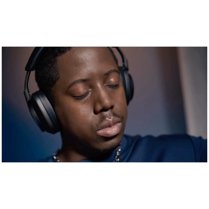 Portrait of a young adult male with closed eyes, wearing headphones, a blue shirt, and a chain against a gradient blue background.