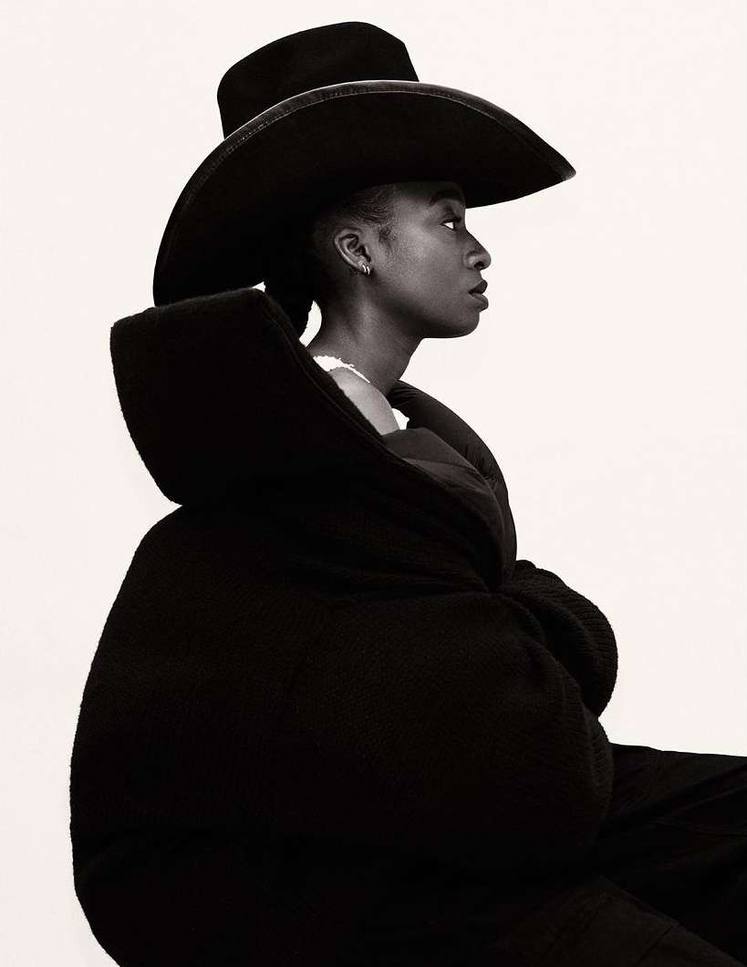 Side-view portrait of a Black woman wearing a wide-brimmed hat and oversized coat against a bright white background. Fashion-editorial.