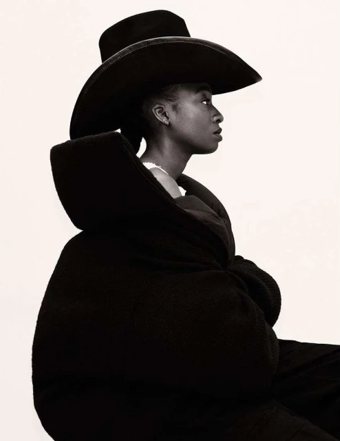 Side-view portrait of a Black woman wearing a wide-brimmed hat and oversized coat against a bright white background. Fashion-editorial.