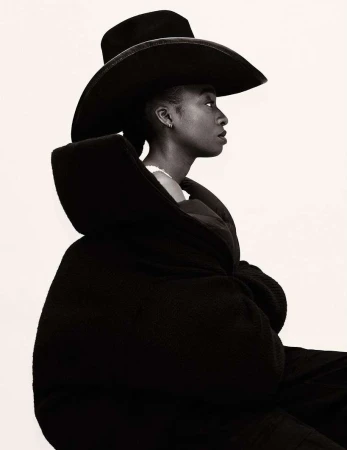 Side-view portrait of a Black woman wearing a wide-brimmed hat and oversized coat against a bright white background. Fashion-editorial.