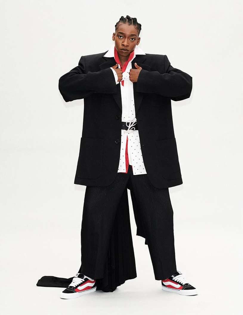 A person in an oversized black suit with white collared shirt and red accents poses in front of a white background in studio lighting.