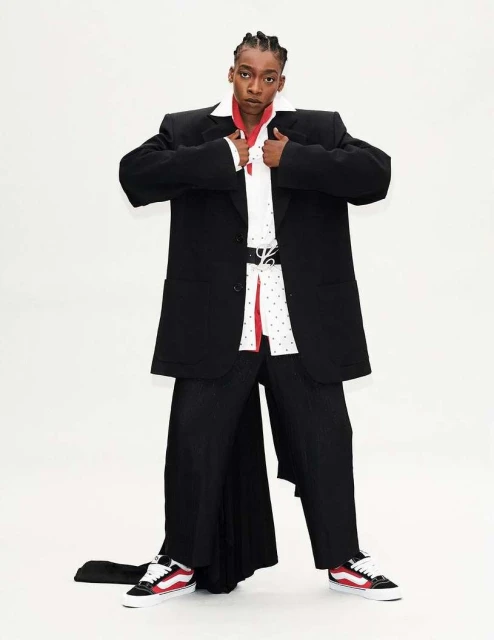 A person in an oversized black suit with white collared shirt and red accents poses in front of a white background in studio lighting.