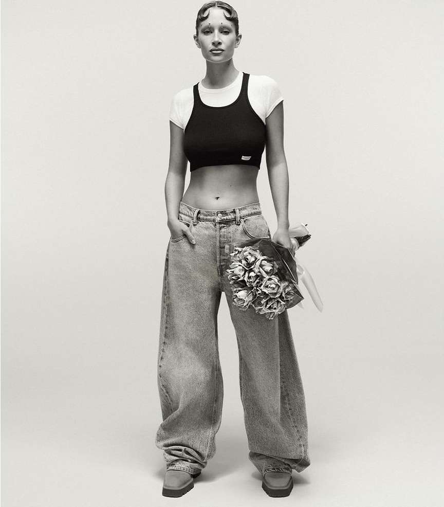 Studio shot of a young woman posing with a bouquet, wearing a black crop top, white t-shirt, baggy jeans, and platform shoes.