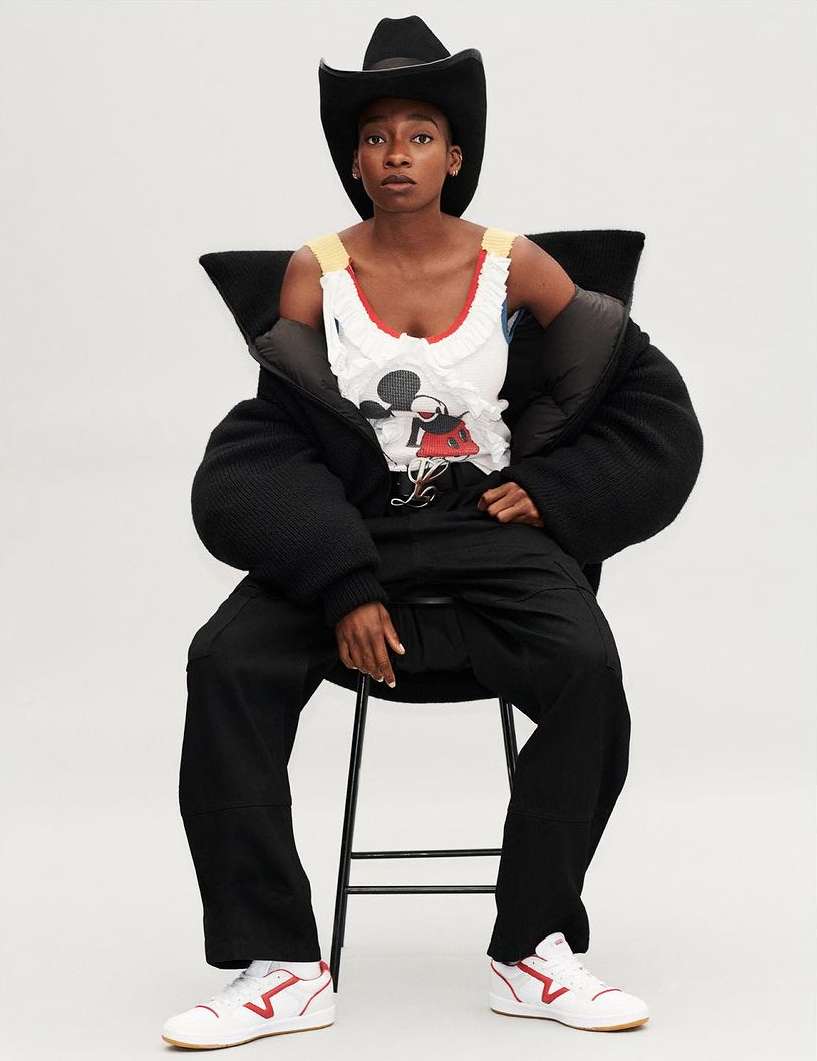Woman in cowboy hat, oversized puffer jacket, and t-shirt with graphic print seated on a chair against a white backdrop.