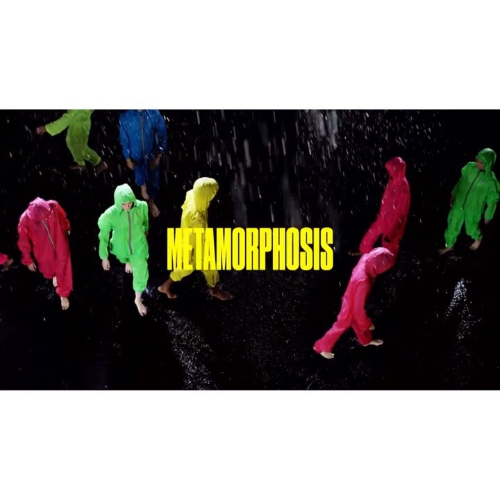 Group of people in colorful hooded jumpsuits walking in rain at night, viewed from above, with "METAMORPHOSIS" text overlay.