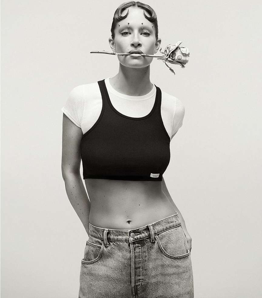 Monochrome portrait of an adult woman posing indoors with a white top and black crop top, holding a rose in her mouth and wearing jeans.