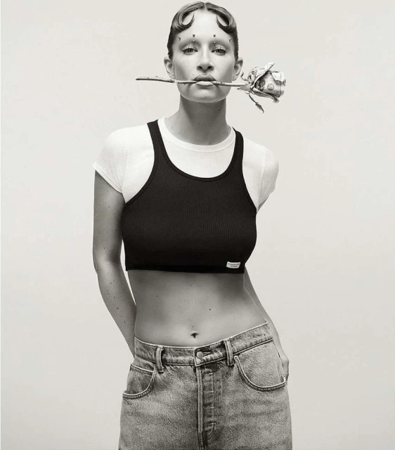 Monochrome portrait of an adult woman posing indoors with a white top and black crop top, holding a rose in her mouth and wearing jeans.