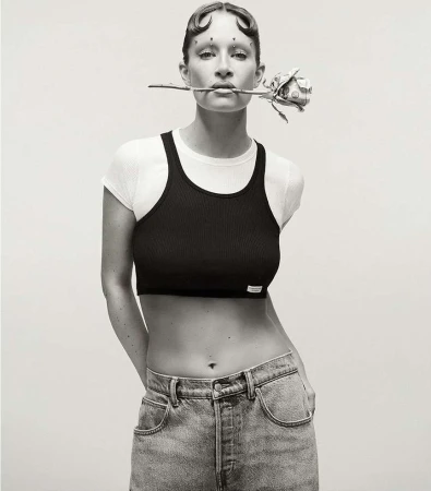 Monochrome portrait of an adult woman posing indoors with a white top and black crop top, holding a rose in her mouth and wearing jeans.