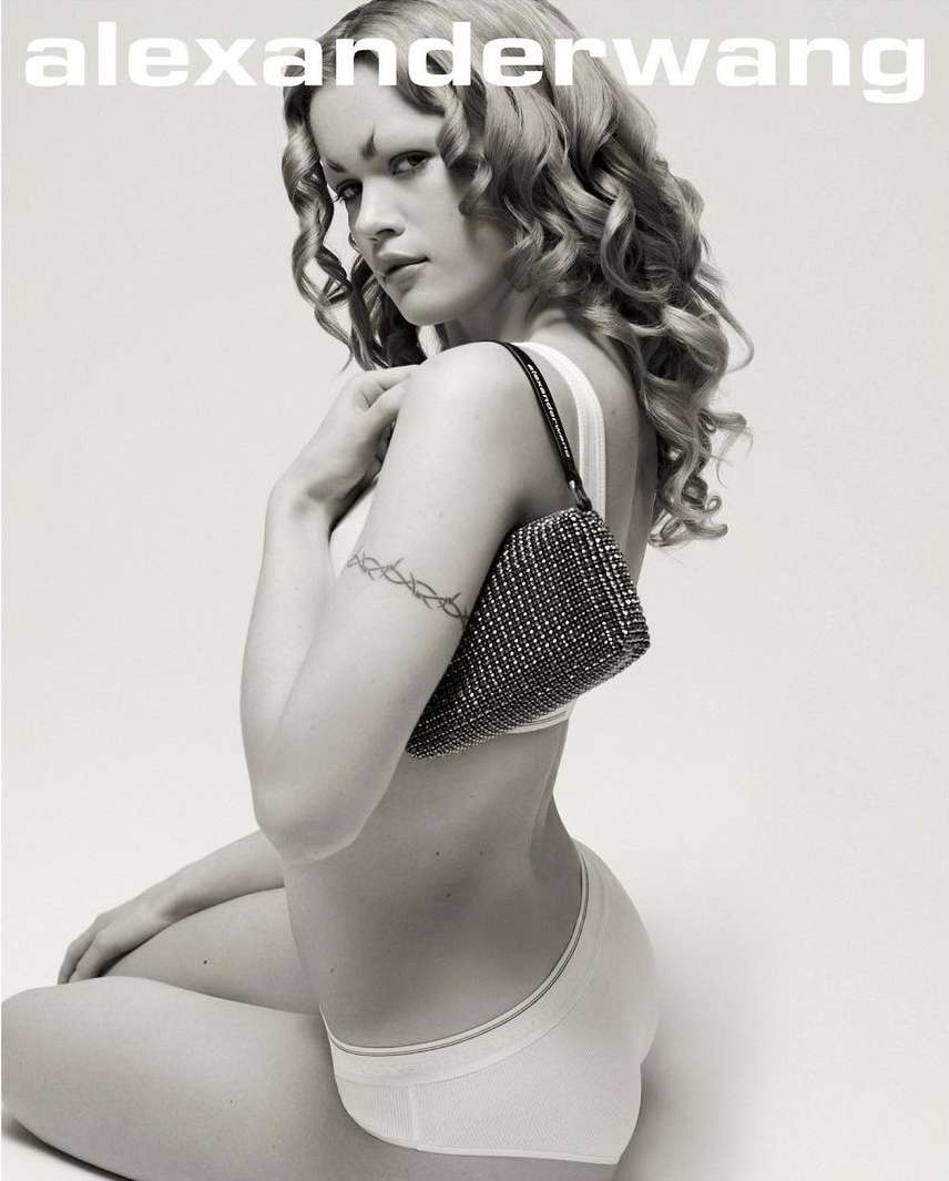 Woman poses with a small beaded purse against a white background. The model is wearing white lingerie and has curly blonde hair.