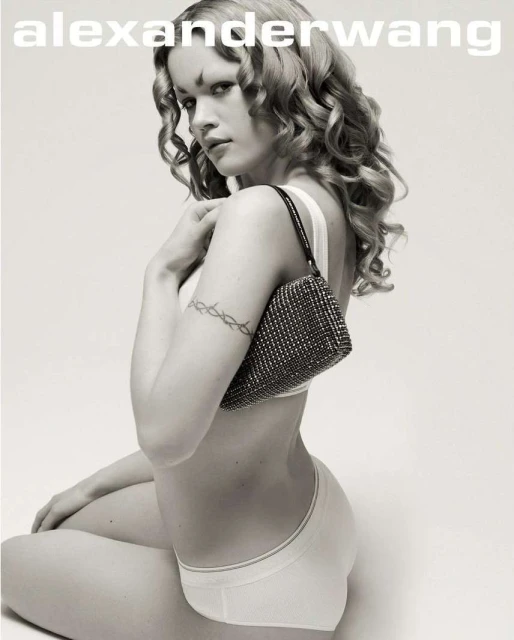 Woman poses with a small beaded purse against a white background. The model is wearing white lingerie and has curly blonde hair.