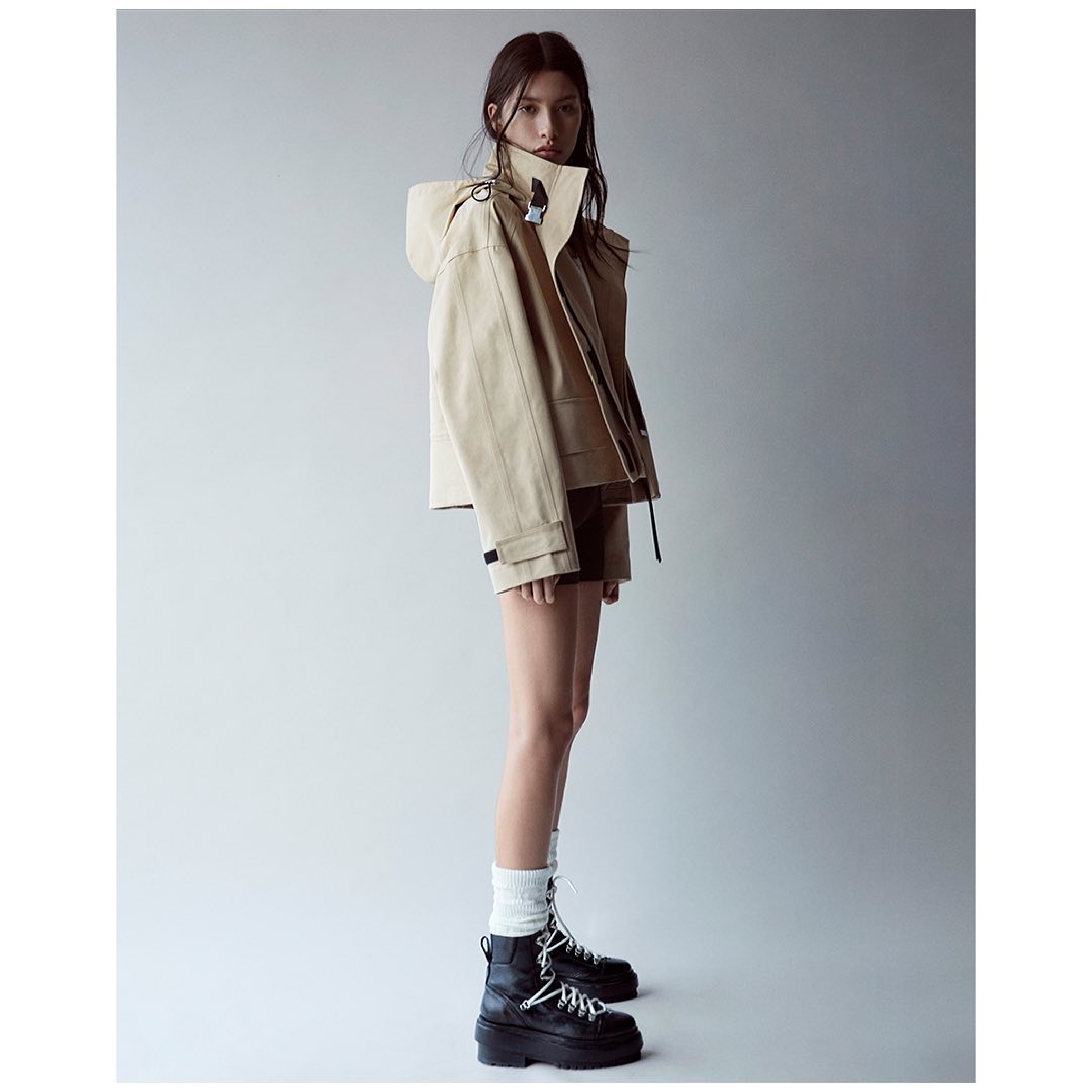 A woman poses in a tan hooded jacket, brown shorts, white socks, and black platform boots against a neutral gray background.