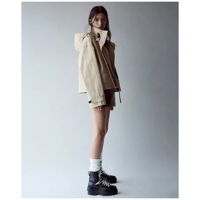 A woman poses in a tan hooded jacket, brown shorts, white socks, and black platform boots against a neutral gray background.
