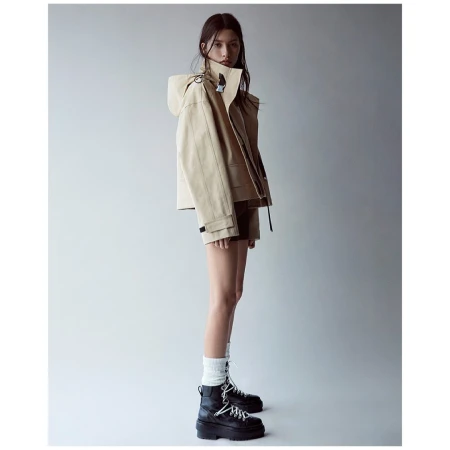 A woman poses in a tan hooded jacket, brown shorts, white socks, and black platform boots against a neutral gray background.