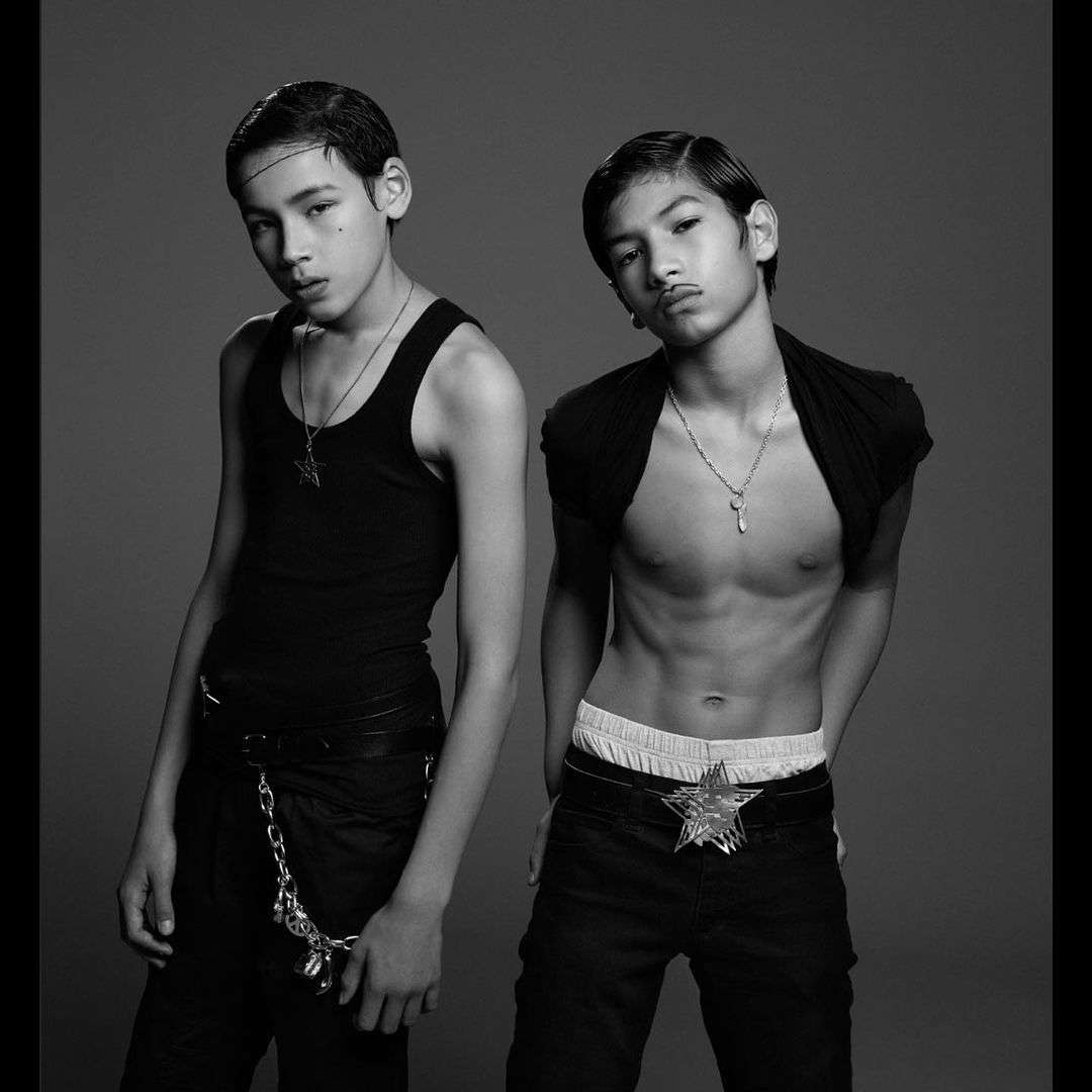 Black and white studio portrait of two adolescent males in dark clothing, one bare-chested with low-slung pants and a star belt buckle.