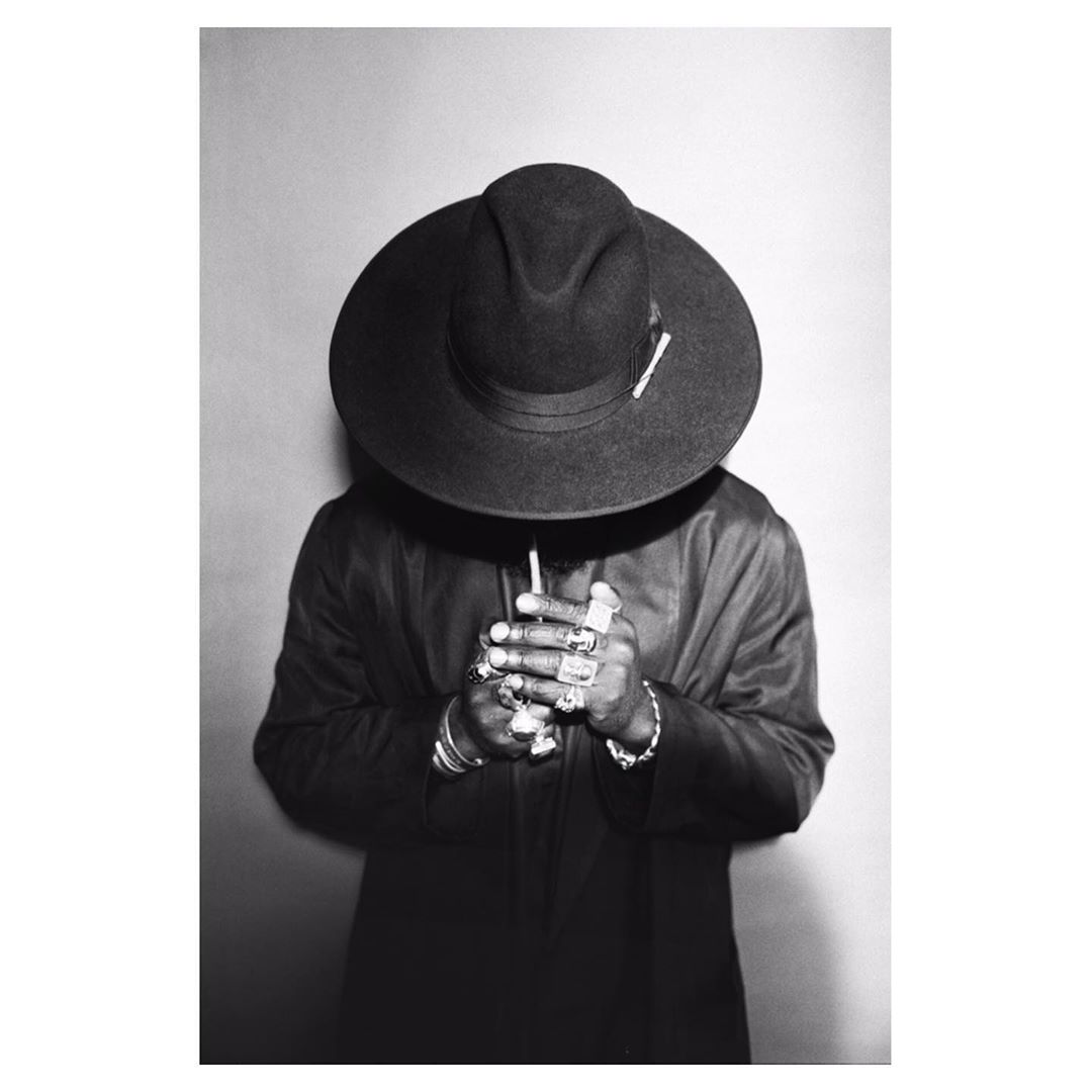 Black and white photo of a person in a wide-brimmed hat and jacket with rings on their fingers, holding a cigarette indoors.