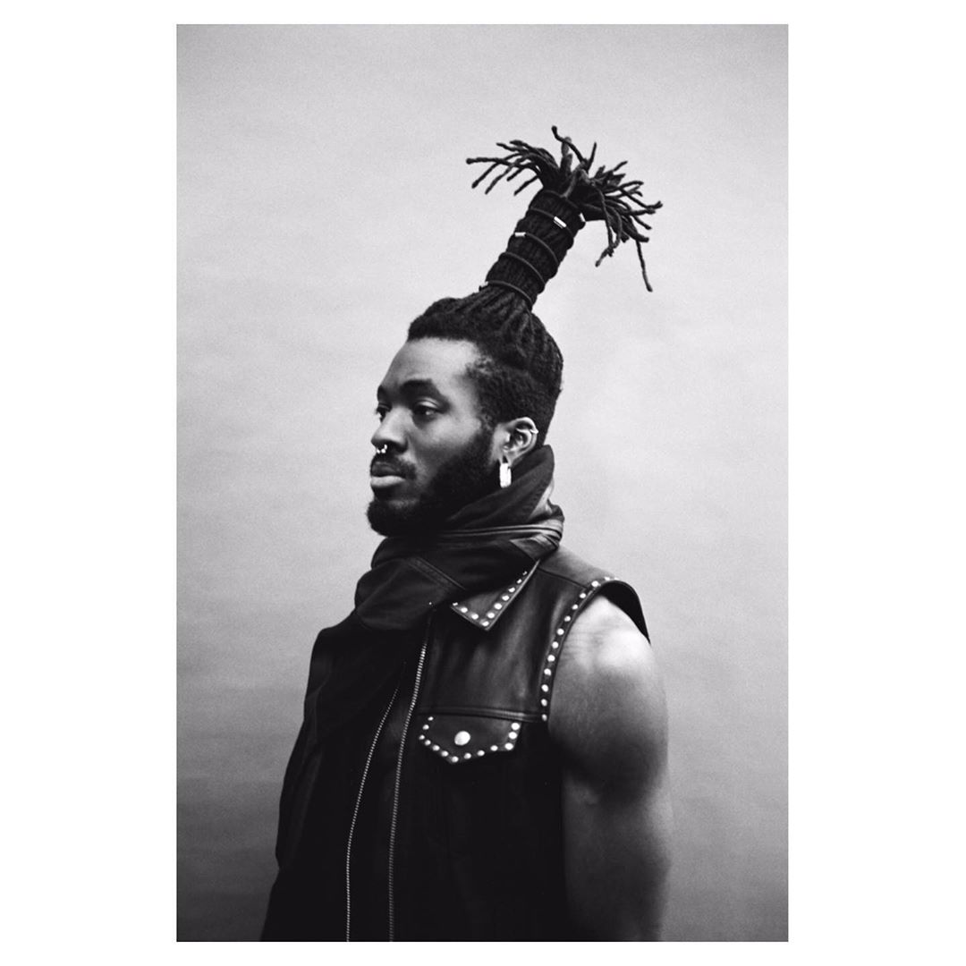 Monochrome portrait of a black man with dreadlocks, wearing a studded vest and scarf, against a plain grey backdrop in a studio setting.