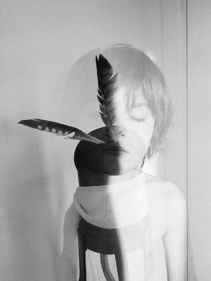 Monochrome double exposure of a person with closed eyes superimposed with a stool and feathers against a textured white wall.