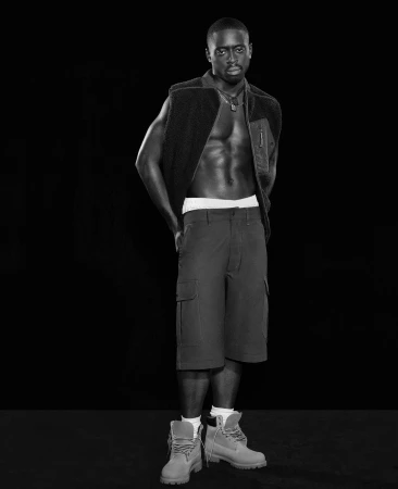 Full-length black and white studio shot of an adult male wearing a vest, shorts, and boots posing against a dark backdrop.