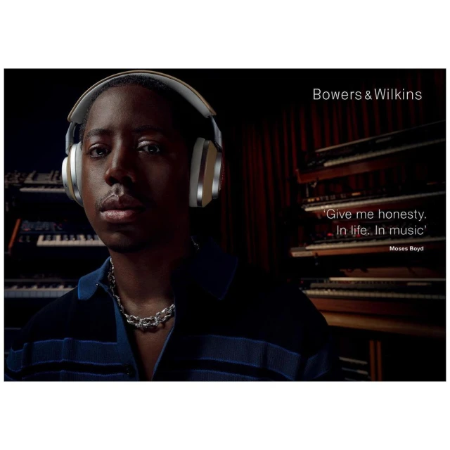 Close-up portrait of an adult male wearing Bowers & Wilkins headphones and a silver chain necklace, with keyboards in the background.