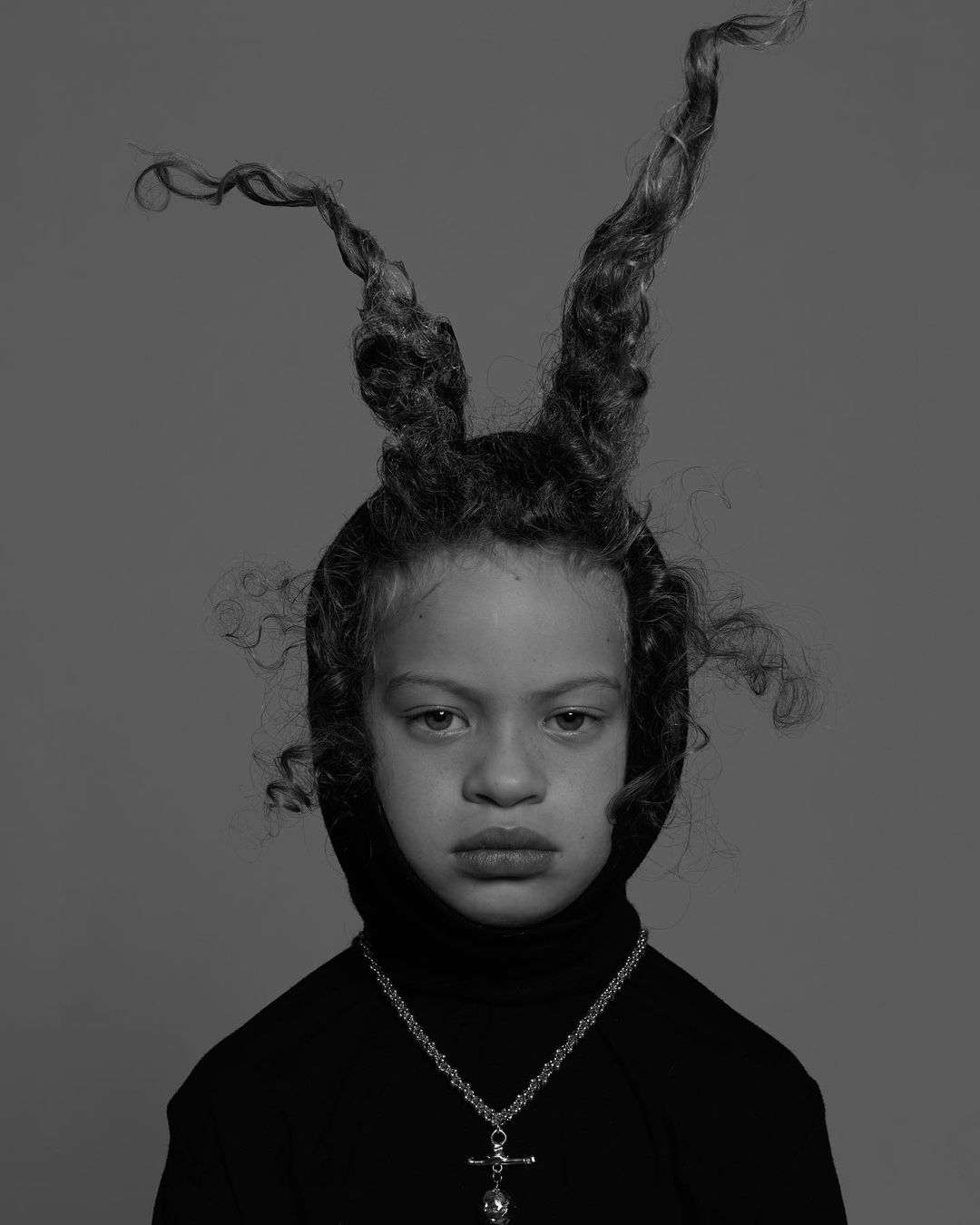 Portrait of a child with styled curly hair resembling horns, wearing a black turtleneck and a cross pendant against a grey background.