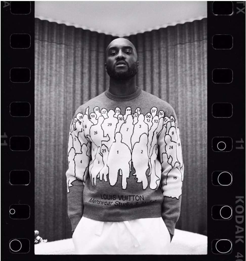 Monochrome portrait of an adult male wearing a printed Louis Vuitton sweater standing indoors with a neutral expression, black-and-white film effect.