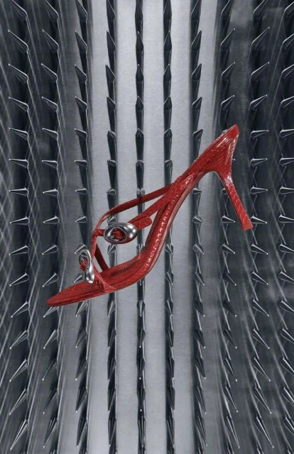 Elevated shot of a glossy red stiletto-heeled sandal against an abstract silver background with metal spike accents and studio lighting.