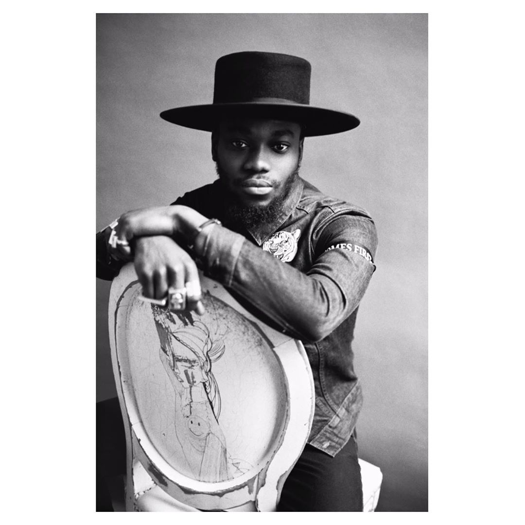 Black and white portrait of a stylish Black man wearing a wide-brimmed hat and denim jacket, seated on an art-covered chair.