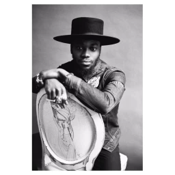 Black man wearing a wide-brimmed black hat and denim shirt leaning over a decorative chair in London by Jamie Morgan