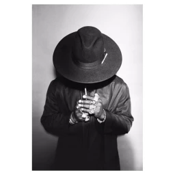 Monochrome portrait of a person wearing a wide-brimmed hat and dark jacket with multiple rings on their hands.