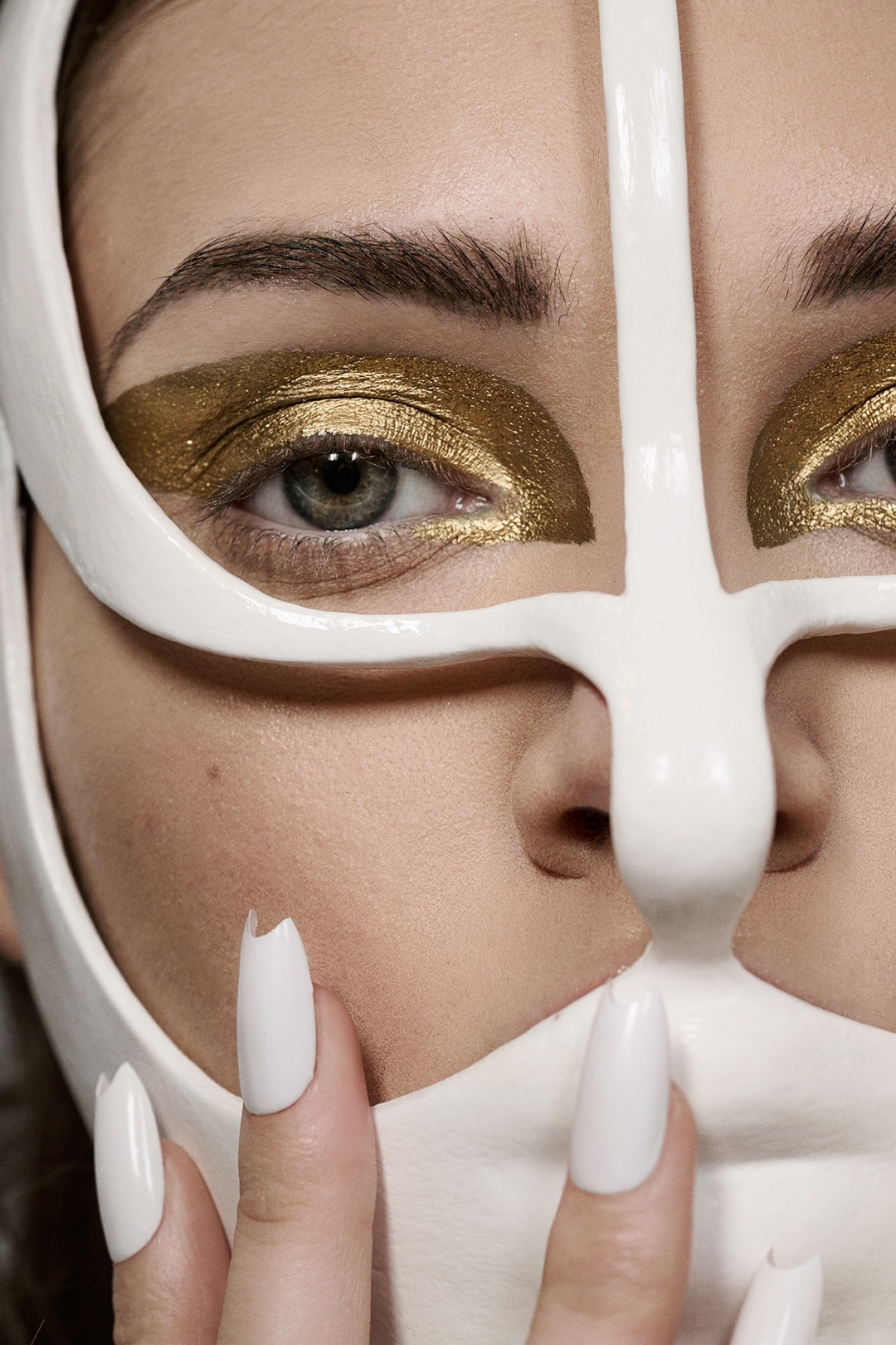 Close-up of a woman's face with bold gold eyeshadow and white mask-like eye ornamentation, showcasing avant-garde beauty concept.