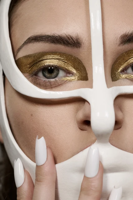 Close-up of a woman's face with bold gold eyeshadow and white mask-like eye ornamentation, showcasing avant-garde beauty concept.