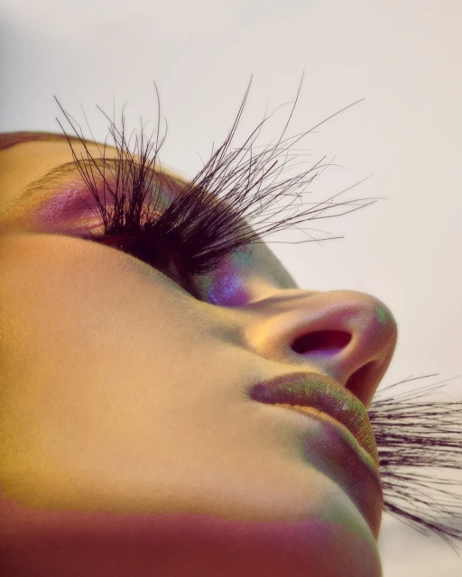 Close-up studio shot of a woman's face angled upward, emphasizing long, dramatic eyelashes and iridescent makeup. Lighting is bright and colorful.