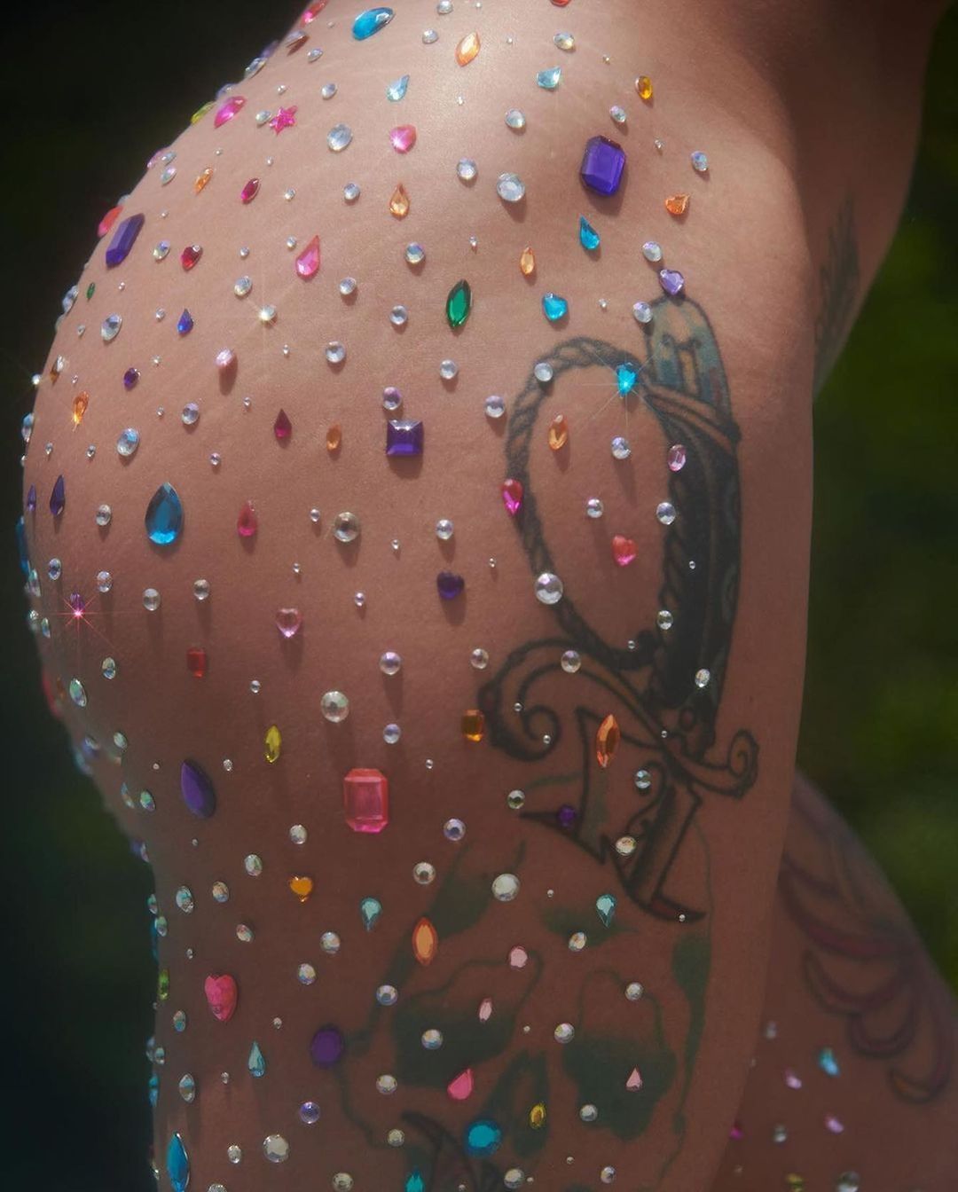 Close-up of an adult's skin adorned with colorful jewels and a tattoo, set against a blurred outdoor background, highlighting body art.