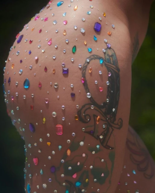 Close-up of an adult's skin adorned with colorful jewels and a tattoo, set against a blurred outdoor background, highlighting body art.