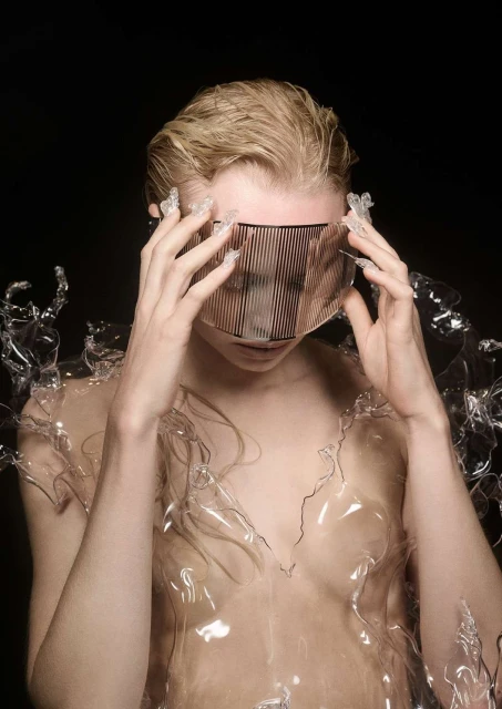 Woman with short blonde hair wearing a translucent visor and plastic garment against a black background in a studio-lit fashion shot.