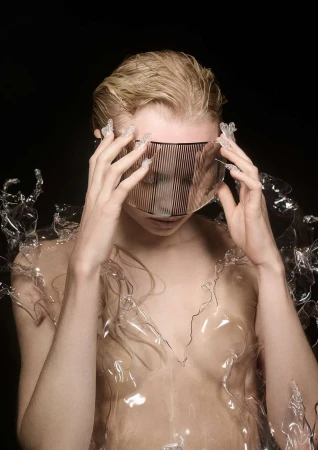 Woman with short blonde hair wearing a translucent visor and plastic garment against a black background in a studio-lit fashion shot.
