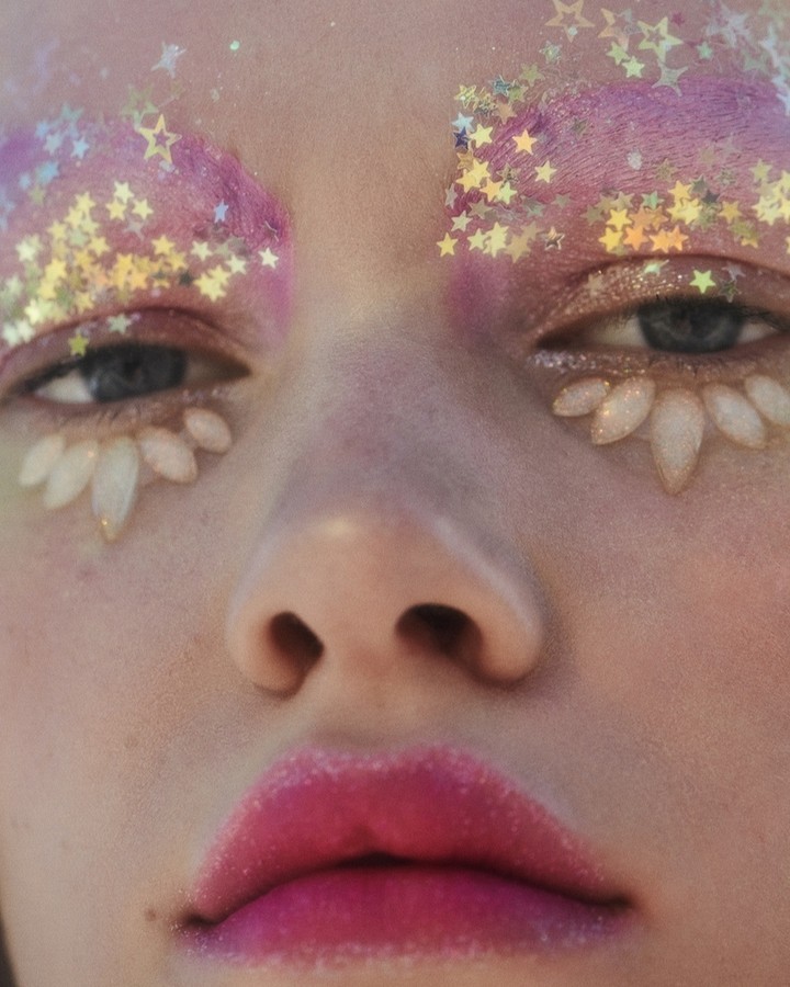 Close-up studio shot of a young adult woman with artistic makeup and glitter around the eyes and pink lipstick, portrait-style.