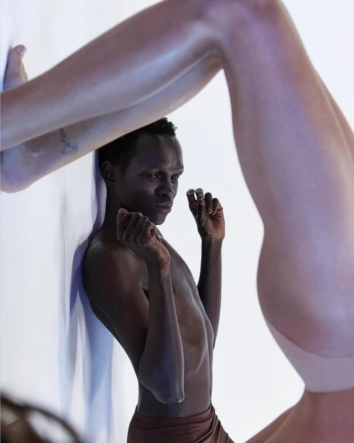 Abstract fashion shot featuring a Black adult male standing near a white wall, with another person's legs framing the scene in soft lighting.