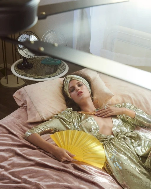 Woman in a sequined jumpsuit reclines on a pink bed with a golden hand fan, wearing a head wrap and jewelry in a stylized indoor fashion shot.