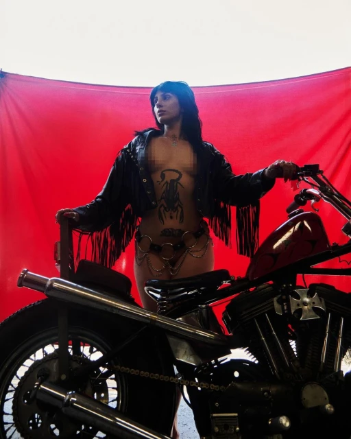 Tattooed woman in leather jacket poses with vintage motorcycle against red backdrop in a studio, exhibiting fashion and automotive elements.