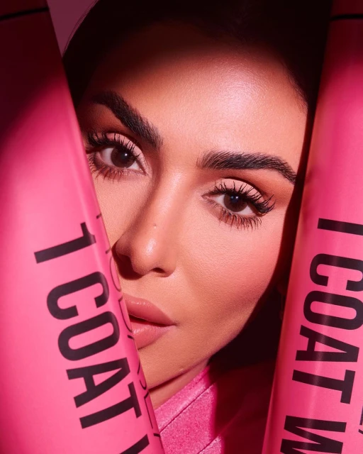 Close-up portrait of an adult female with light skin, holding pink mascara tubes in a beauty advertisement with soft lighting.