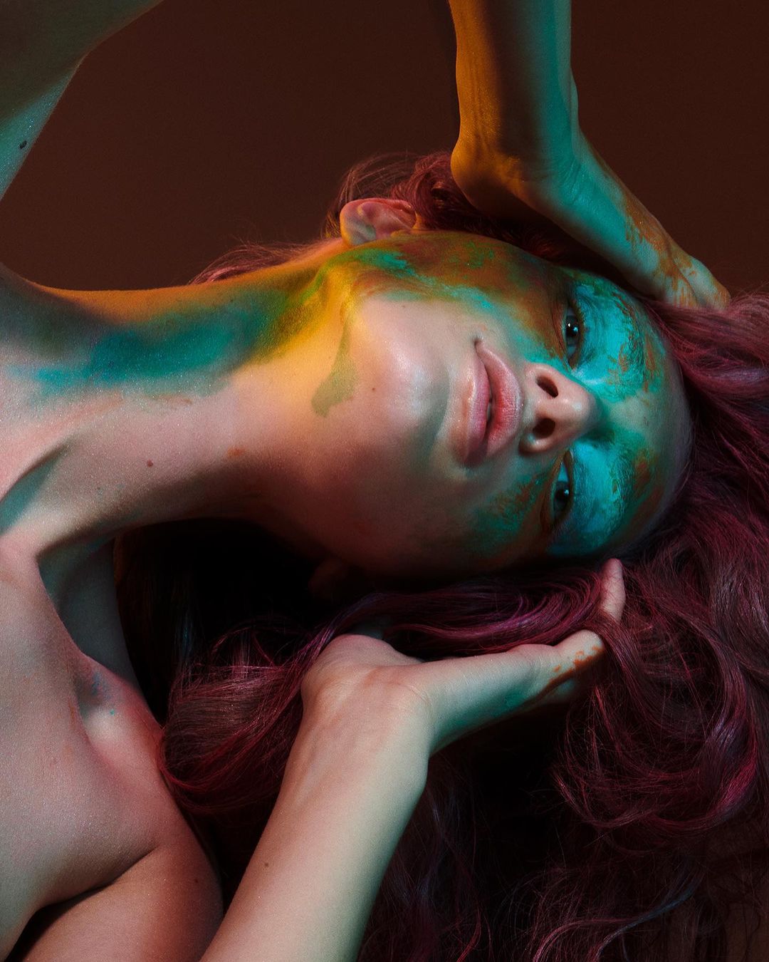 Portrait of a woman with teal and gold body paint and pink hair against a brown background, shot in a photo studio setting.