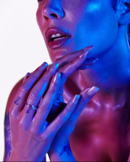 Close-up portrait of a woman's face and hands in blue and pink lighting, showcasing her glossy lips and jewelry.