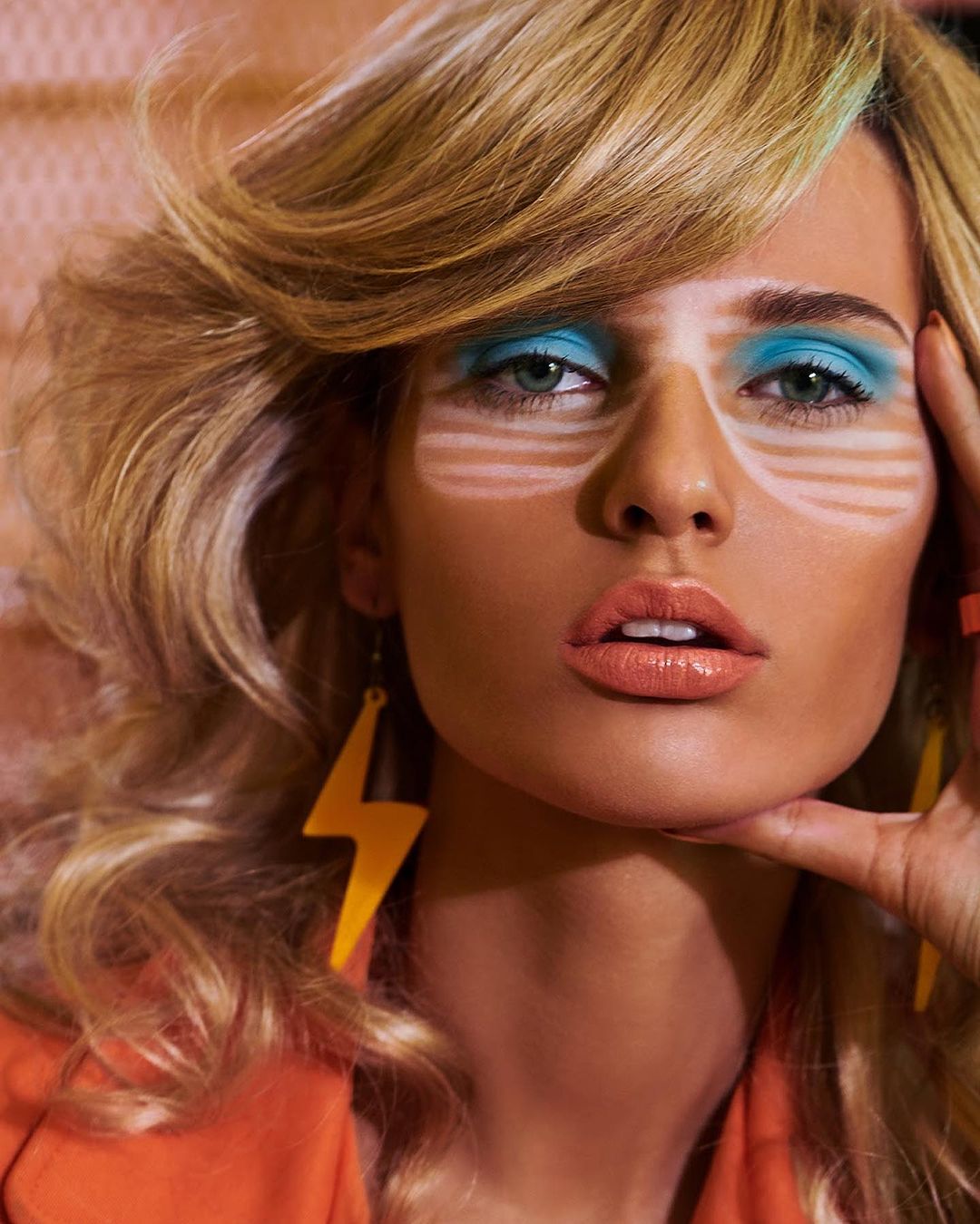 Close-up of a woman with blonde wavy hair and blue makeup posing indoors, wearing orange jacket and a lightning bolt earring.