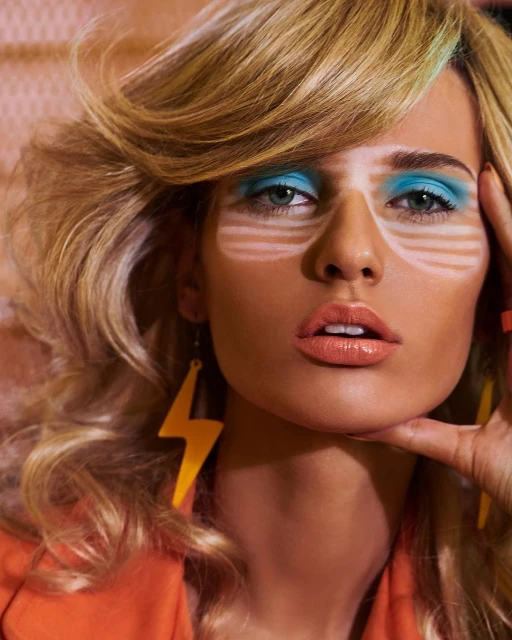 Close-up of a woman with blonde wavy hair and blue makeup posing indoors, wearing orange jacket and a lightning bolt earring.