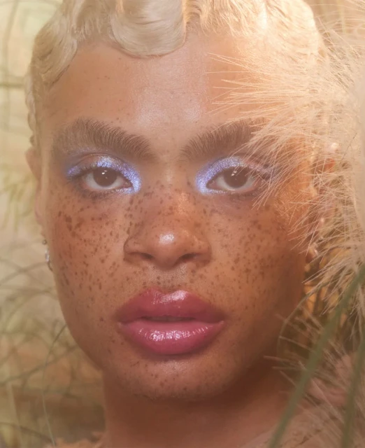Close-up portrait of a fair-skinned adult female with freckles, shimmering light blue eyeshadow, and glossy lips, framed by feathery plants.