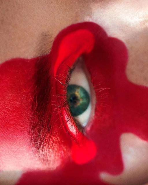 Close-up of a person's eye with green iris surrounded by red paint, creating a surreal and artistic visual.