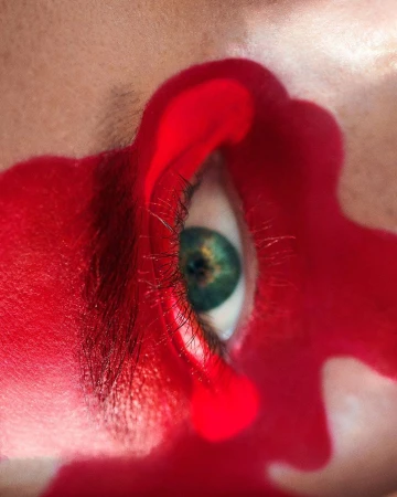 Close-up of a person's eye with green iris surrounded by red paint, creating a surreal and artistic visual.