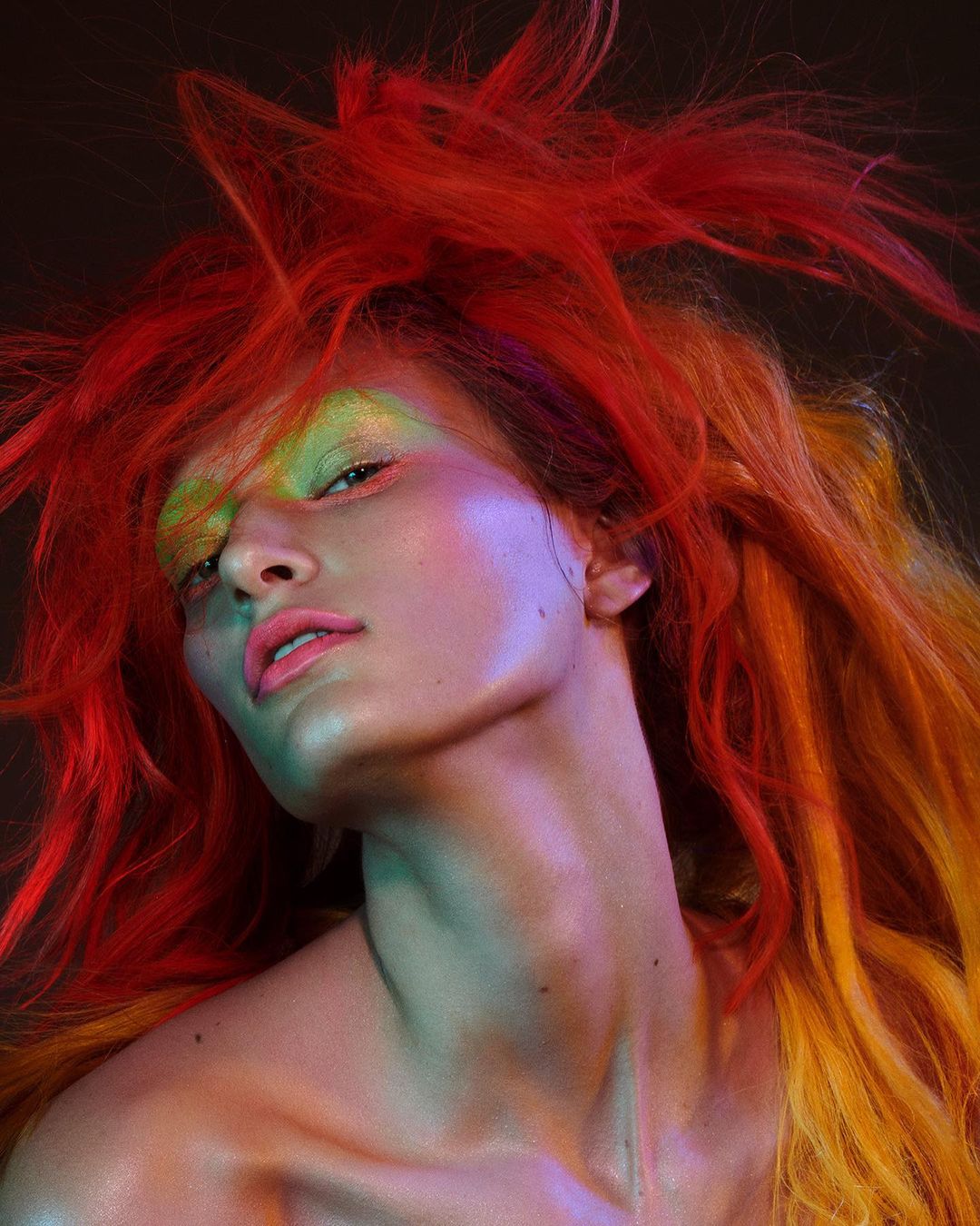 Close-up portrait of a young adult woman with vibrant red and yellow hair, artistic makeup, and a bare shoulder posing against a dark backdrop.
